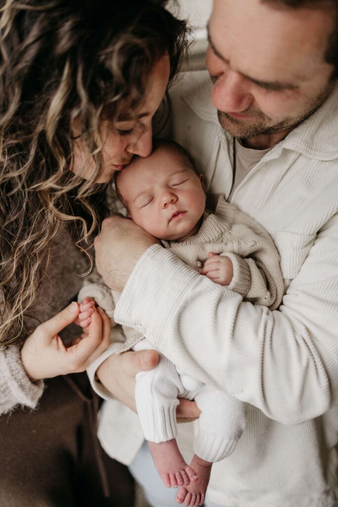 newbornshoot bij papa en mama Baby wordt vastgehouden door papa, mama kust de baby op het hoofd en houdt het handje vast.